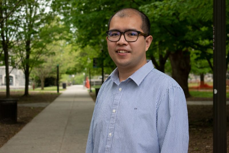 Zichao Yang, graduating with a Ph.D. in economics, focused his dissertation on cryptocurrency Bitcoin. In this photograph, Yang stands along the sidewalk between Pamplin and Williams halls, wearing a striped blue and white shirt. Photo by Steven Mackay.